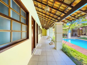 Um corredor coberto com piso de azulejos, portas de madeira e vista para uma piscina.