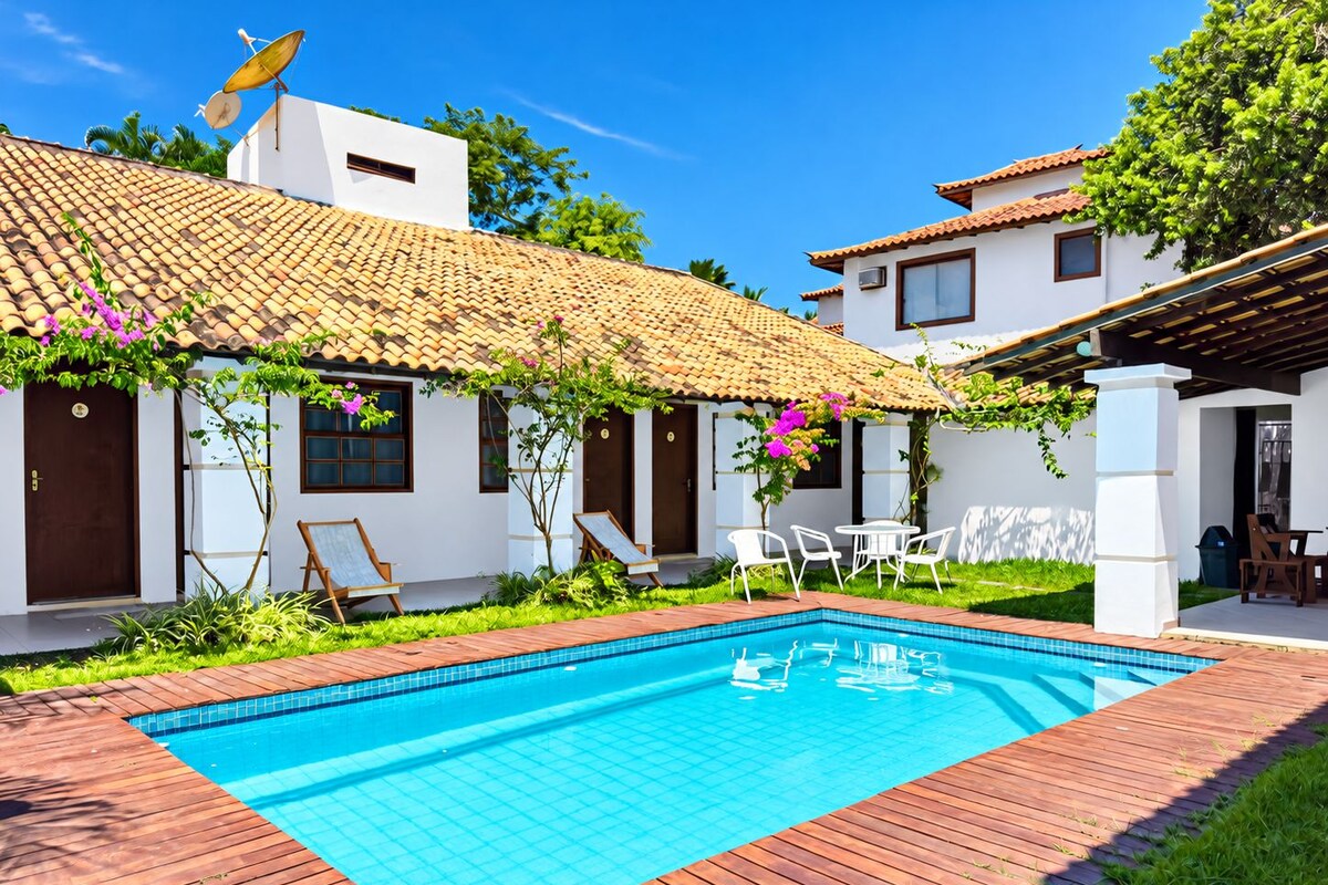 Piscina com deck revestido, cercada por edifícios brancos com telhados azulejados e móveis de exterior.