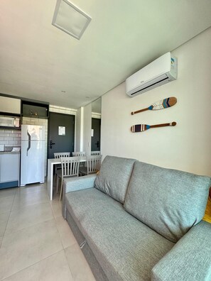 Ein modernes Wohnzimmer mit einem grauen Sofa, einem Essbereich mit weißem Tisch und Stühlen sowie einer Küche mit Kühlschrank und Mikrowelle.