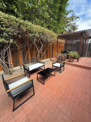 An outdoor patio area with a wooden pergola, two black metal chairs with cushions, and a small table. - Charming Furnished Casita with Private Yard in South Park/Golden Hill (San Diego)
