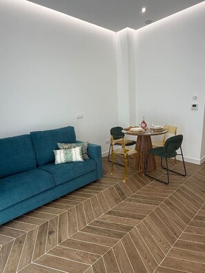 Ein modernes Wohnzimmer mit einem blauen Sofa, einem Esstisch mit Stühlen und einem Holzboden mit Fischgrätmuster.