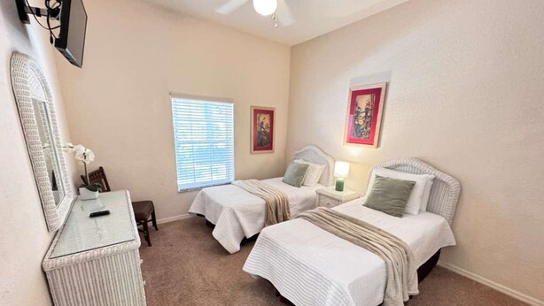 Habitación de hotel con dos camas, un escritorio, una silla, ventilador de techo y obras de arte en las paredes.