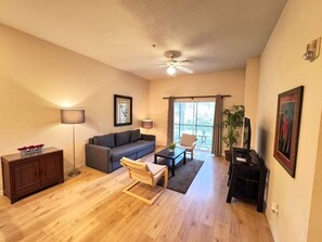 Ein Wohnzimmer mit einer Couch, einem Couchtisch und einem Sessel. Es gibt einen Deckenventilator, eine Lampe und ein Fenster mit Vorhängen.