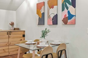 Een eetruimte met een glazen tafel, houten kasten en abstracte wanddecoratie.