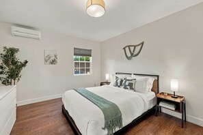 Een slaapkamer met een bed, nachtkastjes, een wandairconditioner, een raam met zonwering en een kunstwerk aan de muur.