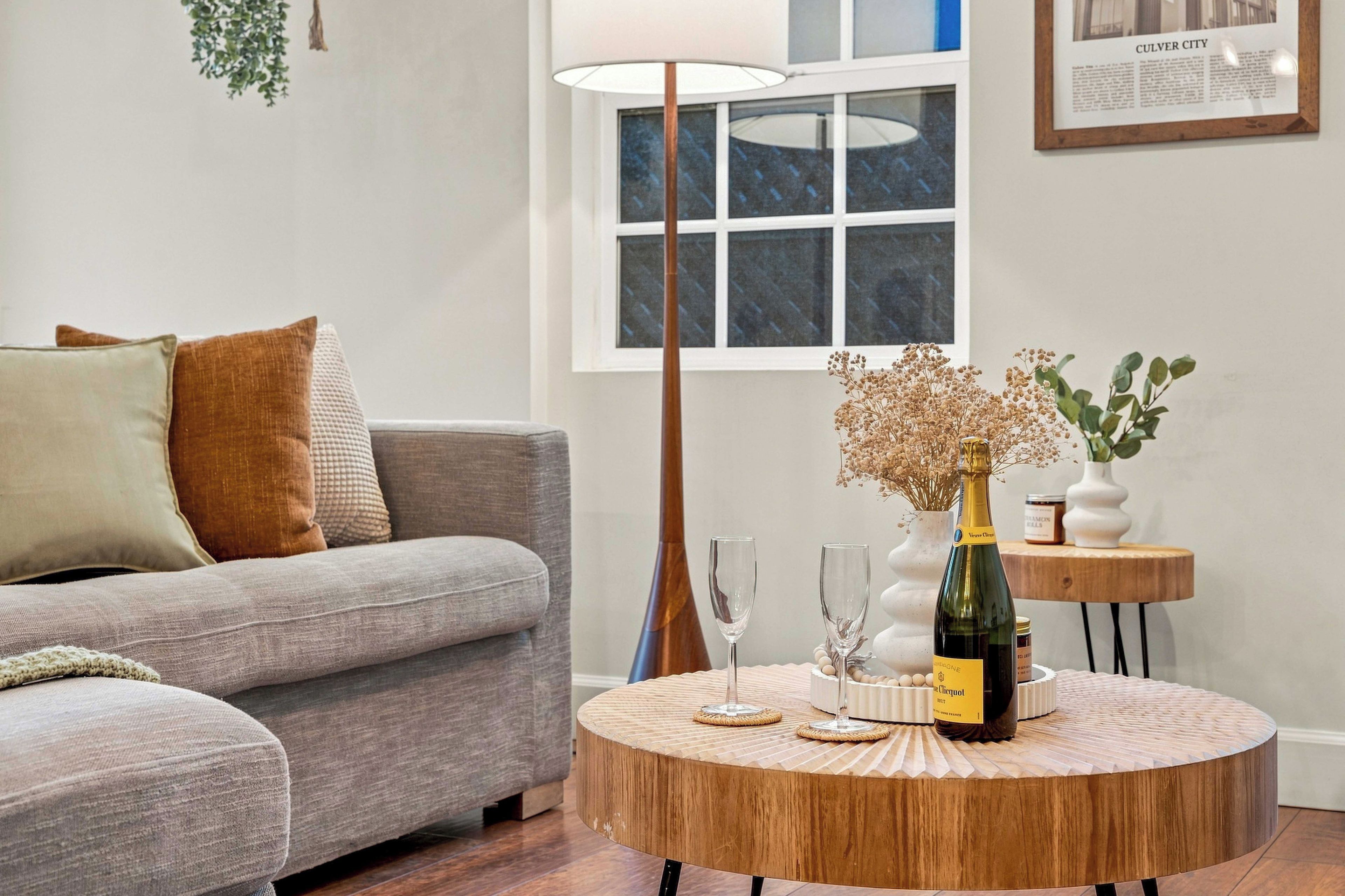 A living room with a grey sofa, a round wooden coffee table, a bottle of champagne, two wine glasses, and a small side table with a vase of dried flowers.