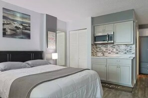 Quarto com cama, mesa de cabeceira, TV na parede e cozinha compacta com armários e micro-ondas.