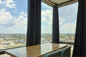 A room with a large window offering a view of a cityscape, a wooden table, and a chair. - Top floor corner oceanview panoramic studio w/pool (Daytona Beach)
