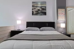 A bedroom with a bed, two bedside tables, a nightstand, a wall-mounted artwork, and a poster. - Top floor corner oceanview panoramic studio w/pool (Daytona Beach)