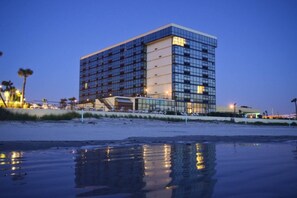 A modern hotel with a reflective glass facade, situated by a beach with a sandy shoreline. - Top floor corner oceanview panoramic studio w/pool (Daytona Beach)