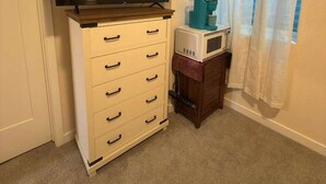 Ein Zimmer mit Fernseher auf einem Schrank, einer Mikrowelle auf einem kleinen Tisch und einem Fenster mit Vorhängen.