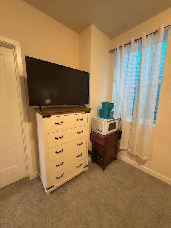 A room with a television on a stand, a microwave on a small table, and a window with curtains.
