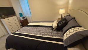 Un dormitorio con cama, cómoda, televisor, lámpara y ventana con cortinas.