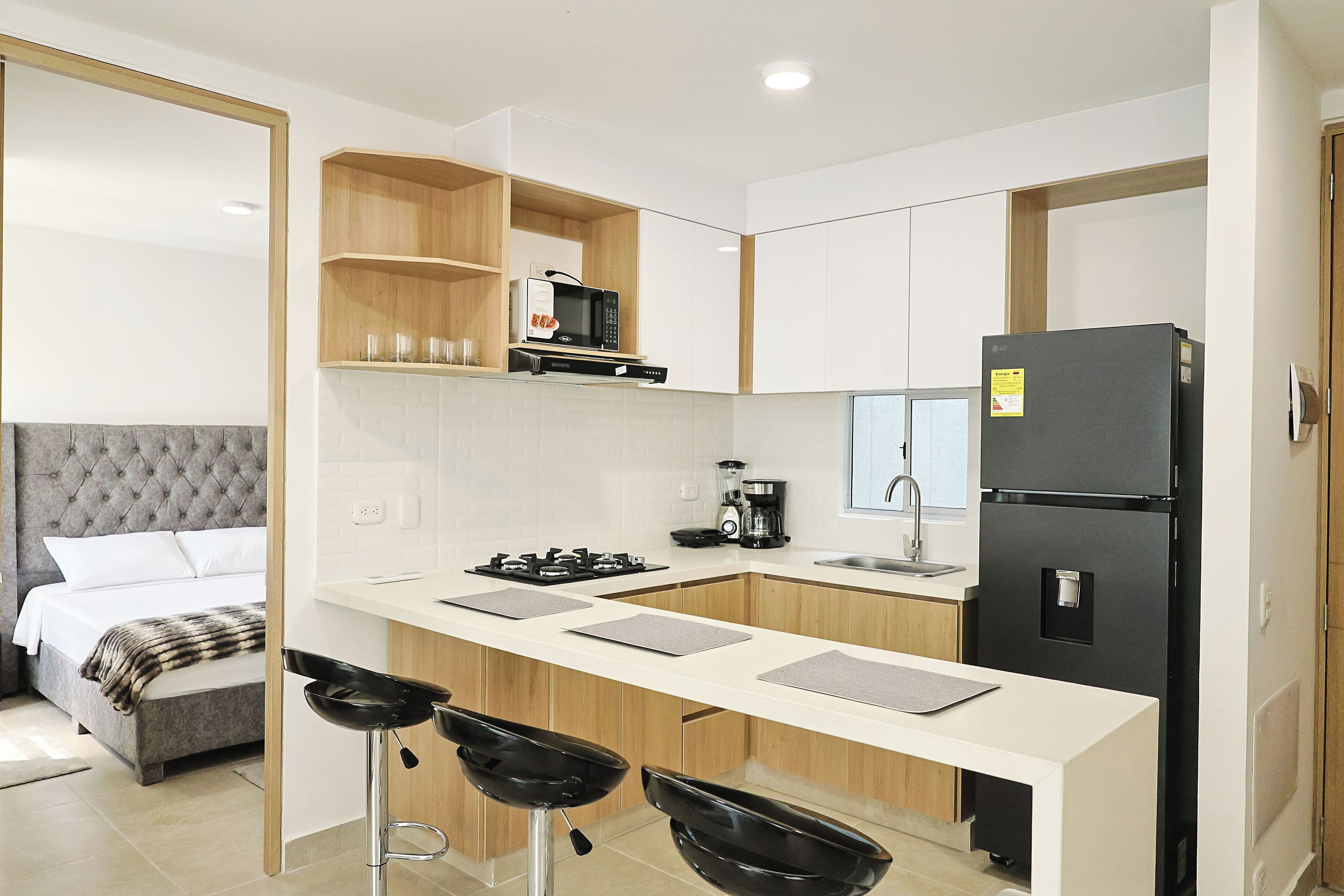 A compact kitchen with a microwave, stove, and refrigerator. There is a bar stool and a bed with a grey headboard in the adjacent room.