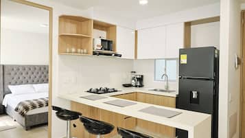 A compact kitchen with a microwave, stove, and refrigerator. There is a bar stool and a bed with a grey headboard in the adjacent room.
