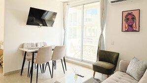Uma sala de estar moderna com televisão de tela plana na parede, um espaço de jantar com mesa e cadeiras, e uma grande janela com cortinas.