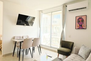 Una sala de estar moderna con un televisor de pantalla plana montado en la pared, una zona de comedor con mesa y sillas, y un ventanal con cortinas.