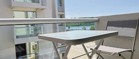 A balcony with a glass table and a chair, overlooking a modern building and a body of water.