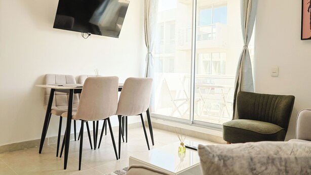 Uma sala de estar moderna com televisão de tela plana na parede, um espaço de jantar com mesa e cadeiras, e uma varanda com vista para edifícios.
