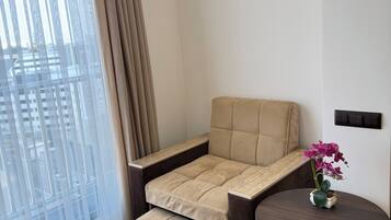 Uma sala de estar moderna com um sofá-cama, uma pequena mesa redonda com um vaso de flores e vista para a cidade através de amplas janelas.