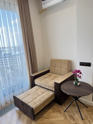 A modern living room with a convertible sofa, a small round table with a vase of flowers, and a view of the city through large windows. - Seoul Riverside apartment (Tashkent)