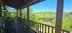 A wooden deck with outdoor seating, overlooking a forested landscape and a bridge.