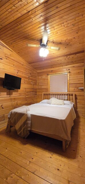 A wooden cabin room with a bed, a ceiling fan, a television, and a window with blinds. - Scenic Riverview Cabin For Fishermen/Outdoorsmen (Buffalo Valley)