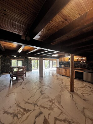 A spacious room with a stone wall, wooden ceiling beams, a dining table with chairs, and a kitchen area with wooden cabinets and appliances.