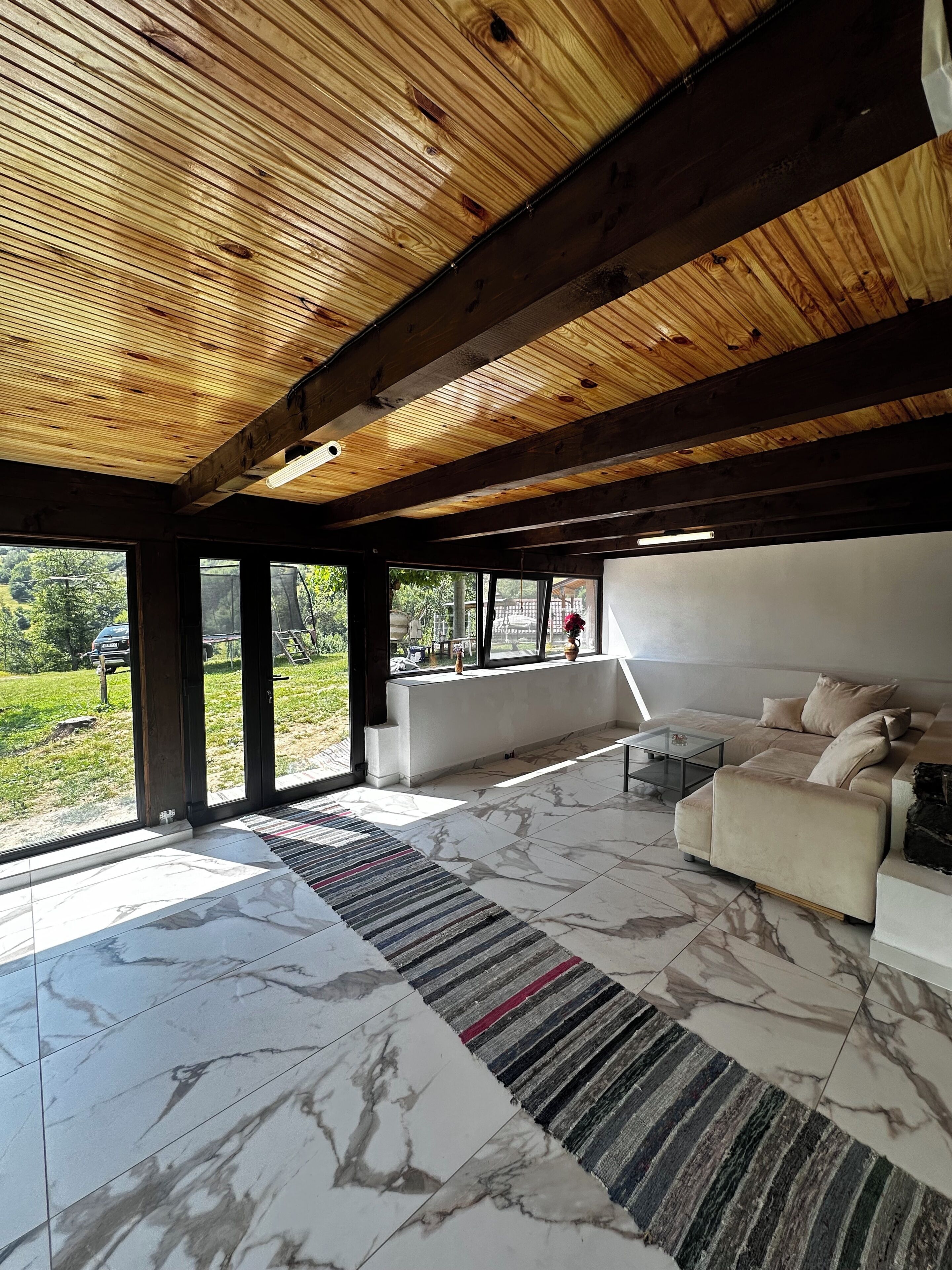 Un vaste espace de vie doté d’un plafond en bois, de grandes fenêtres, d’un canapé beige et d’un sol en marbre.