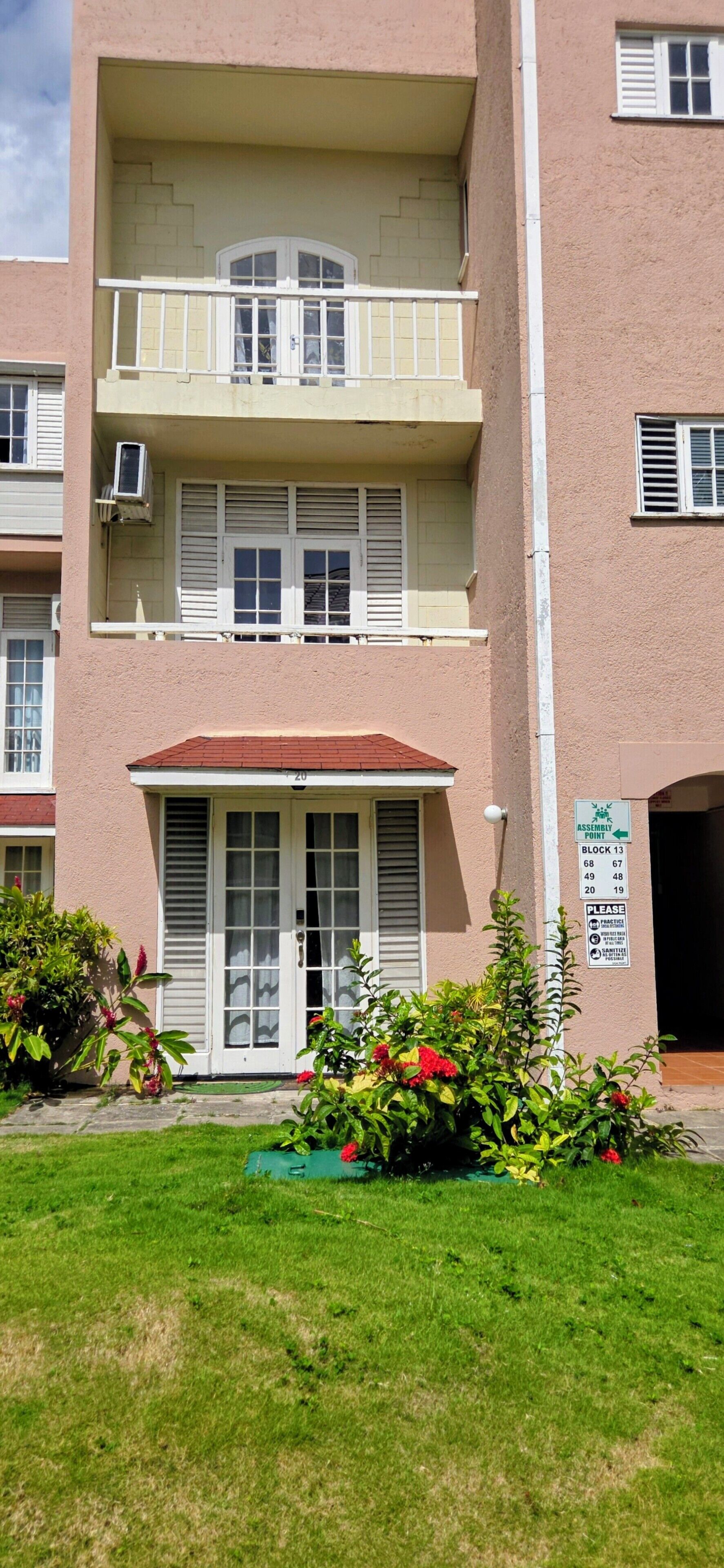 A multi-story building with a central entrance, a balcony on the second floor, and a sign indicating an assembly point.