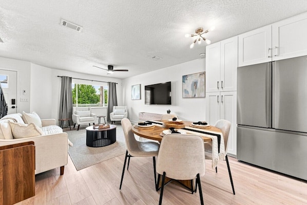 A modern kitchen with a stainless steel refrigerator, a dining area with a wooden table and chairs, and a living room with a sofa and a TV.