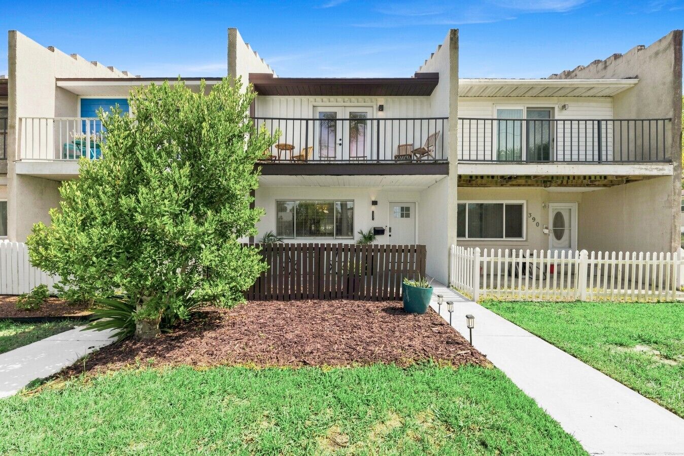 A two-story residential building with a fenced front yard, a small tree, and a pathway leading to the entrance.