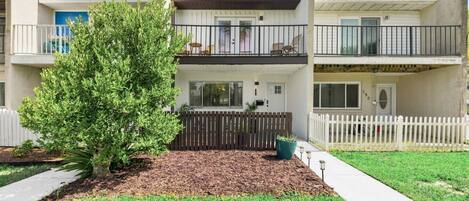 A two-story residential building with a fenced front yard, a small tree, and a pathway leading to the entrance.