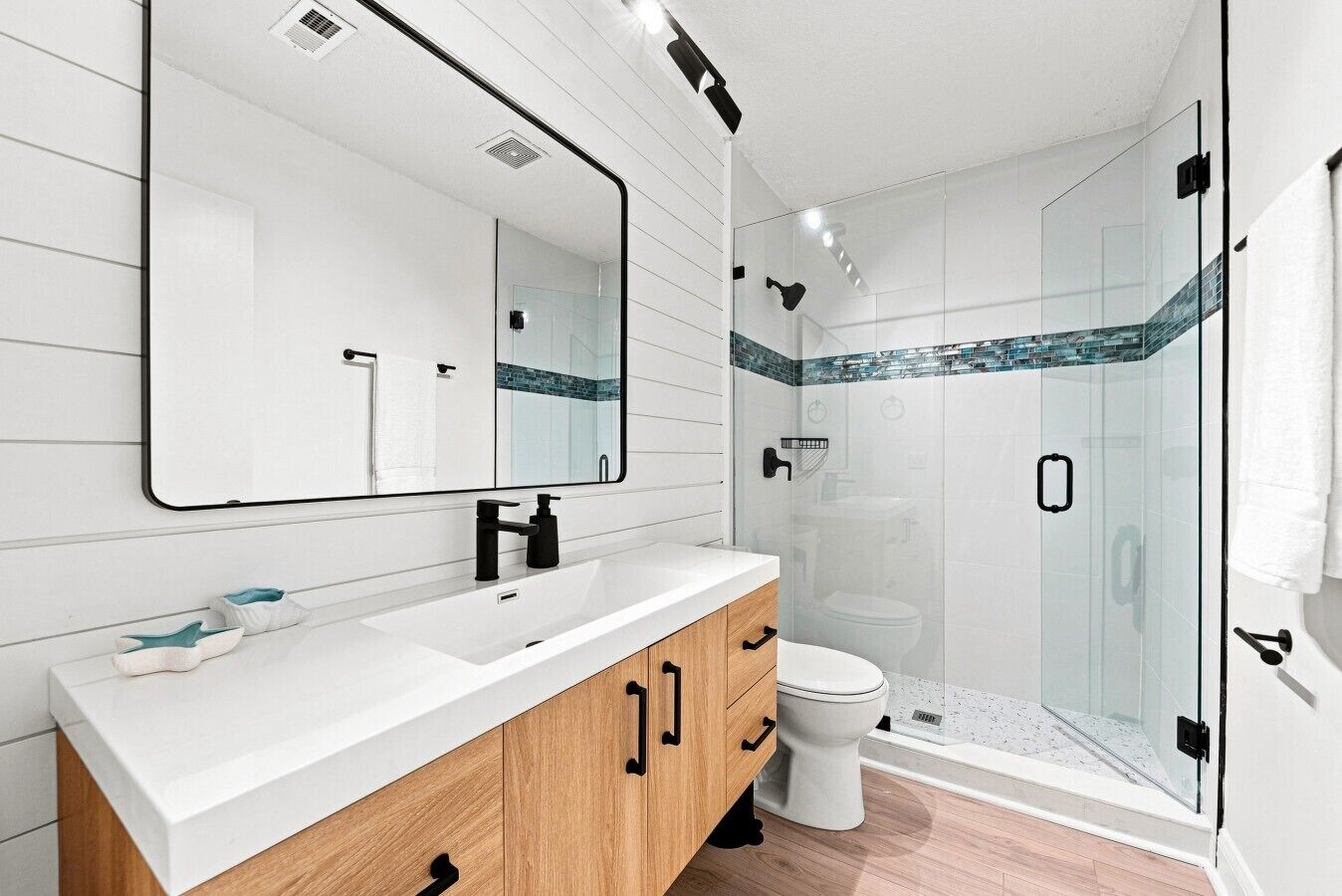 A modern bathroom with a large mirror, white vanity, wooden cabinet, white toilet, and a glass-enclosed shower area.