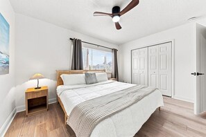 A bedroom with a bed, bedside table, lamp, ceiling fan, window with curtains, and a door.