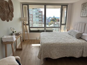 Een slaapkamer met een groot raam, een bed met een beddegoed met patroon, een nachtkastje met handdoeken en een lamp.
