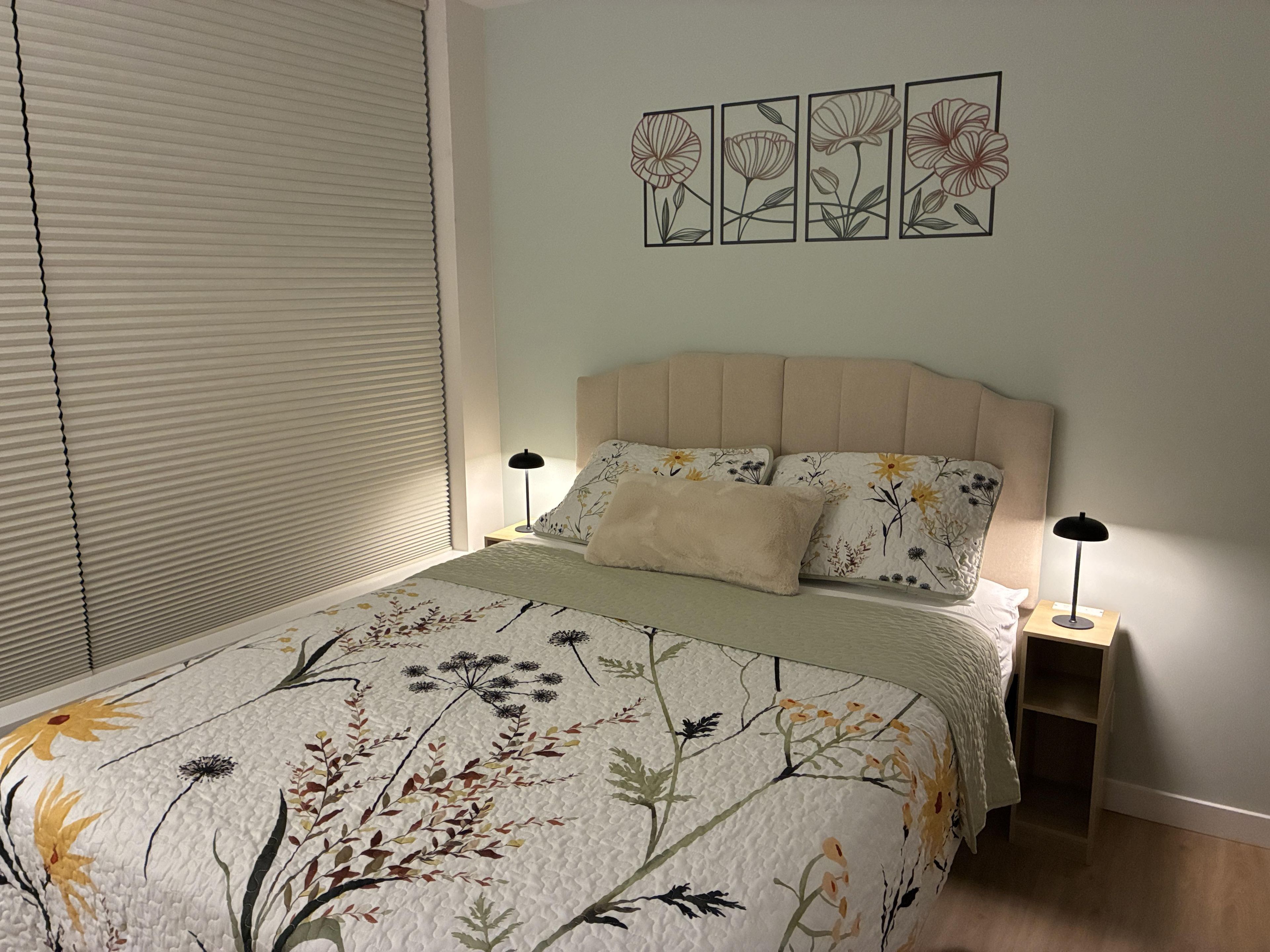 A bedroom with a bed, a nightstand, a wall-mounted artwork, and a window with blinds.