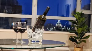 Uma mesa de vidro com duas taças de vinho e uma garrafa, um vaso de planta e vista para o horizonte noturno.