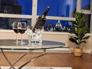 Ein Glastisch mit zwei Weingläsern und einer Weinflasche, ein Topfpflanze und Blick auf eine nächtliche Skyline.