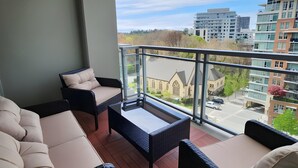 Uma varanda com um sofá, uma mesa pequena e uma cadeira, com vista para um horizonte urbano com edifícios modernos.