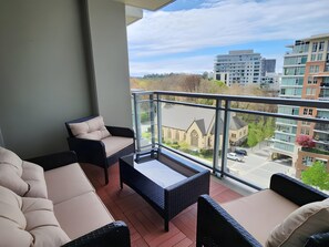 Ein Balkon mit einer Couch, einem kleinen Tisch und einem Stuhl, mit Blick auf eine Skyline moderner Gebäude.