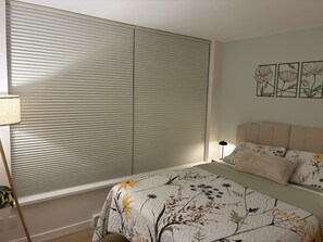 Een slaapkamer met een bed, een nachtkastje, een lamp en een raam met jaloezieën.