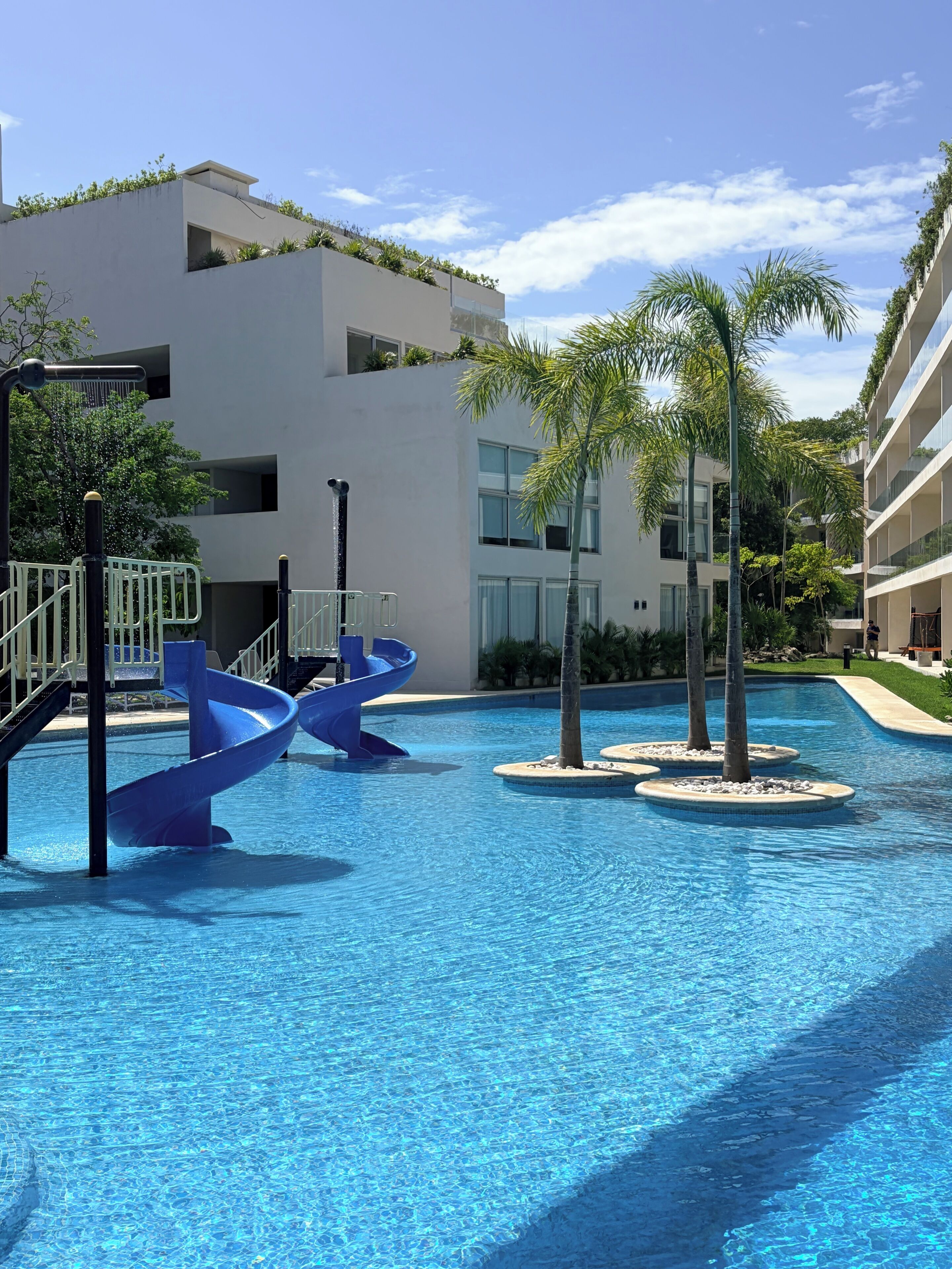 Una piscina con un tobogán azul, rodeada de edificios modernos y palmeras.