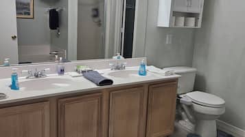A bathroom with a double sink vanity, a toilet, a shower, and a mirror with lights.