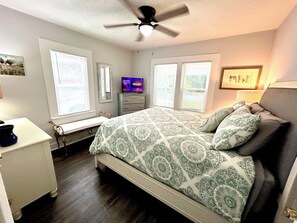 Una camera da letto con un letto, una televisione, una ventola a soffitto e due finestre con tende.