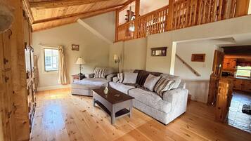A spacious living room with a wooden ceiling, a large sofa set, a coffee table, and a wooden staircase leading to an upper level.