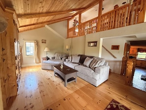A spacious living room with a wooden ceiling, a large sofa set, a coffee table, and a wooden staircase leading to an upper level.