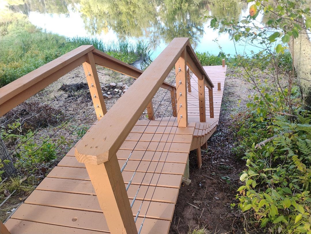Un puente de madera con baranda metálica que conduce a una plataforma de madera sobre un cuerpo de agua.