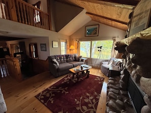 A cozy living room with a sofa, a coffee table, and a rug. There is a telescope on a tripod, a window with curtains, and a stone wall.