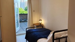 A bedroom with a bed, a bedside table with a lamp, a sliding glass door leading to an outdoor area, and a view of buildings and greenery outside. - Modern Bright Riverside Stay (London)